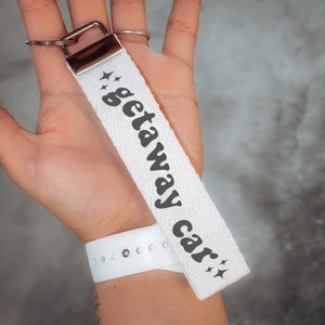 May include: White canvas keychain with the text "getaway car" in black retro font with stars on either side of the text.