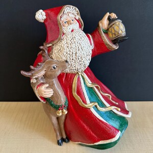 May include: A ceramic Santa Claus figurine in a red coat with gold trim, holding a lantern and a reindeer. The figurine has a white beard and is decorated with green and gold accents. A festive Christmas decoration.