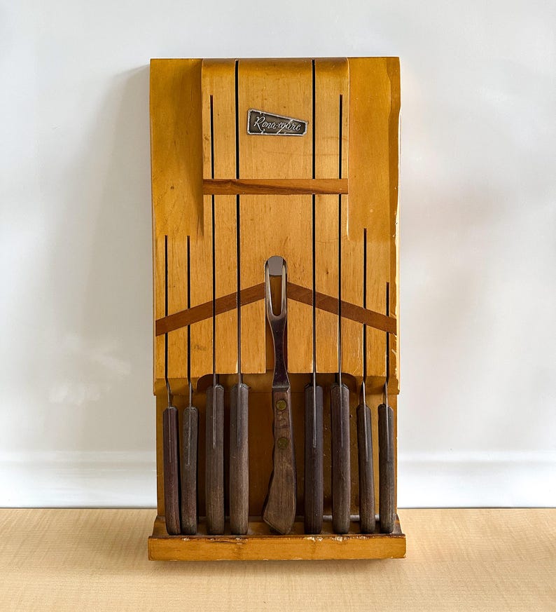 May include: A wooden knife block with a set of knives and a carving fork. The block is light brown and has the word "Ronaglo" printed on it.