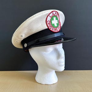 May include: A vintage white and black Crossing Guard hat with the text "L.A.P.D. CROSSING GUARD" on a circular patch. The hat has a black brim and band, and is displayed on a white mannequin head.