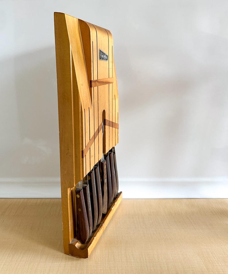May include: A wooden knife block with a light brown finish and a black label that says "Waring-Rex" holds a set of knives with brown handles.