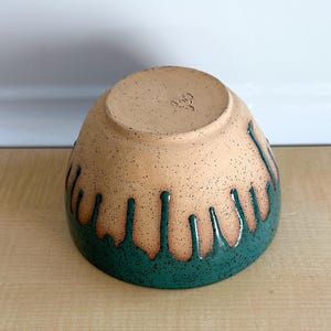 May include: A handmade ceramic bowl with a speckled tan exterior and a teal green drip glaze design. The bowl is inverted, revealing the unglazed base. The bowl is on a light brown surface.
