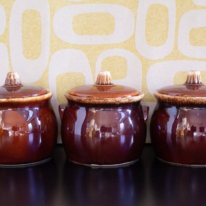 May include: Three brown glazed ceramic casserole dishes with lids. The dishes are round and have a slight curve to them. The lids are slightly domed.