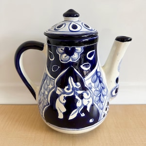 May include: A ceramic teapot featuring a white base with hand-painted deep blue floral and leaf patterns. The teapot includes a matching lid with a knob, a curved handle, and a spout, all decorated with intricate blue designs on a white background.
