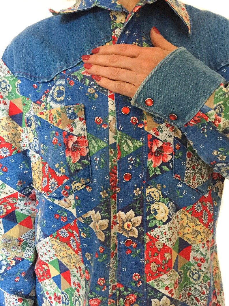 May include: A blue denim shirt with a patchwork design featuring floral patterns in red, yellow, and green. The shirt has a pointed collar and a button-up front.