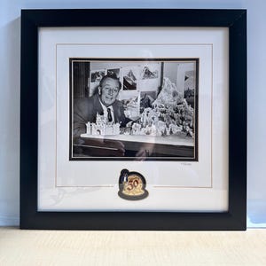 May include: A black and white framed photograph of Walt Disney working on a model of the Matterhorn mountain and a castle. The white matting features a gold-trimmed Celebrating 50 Years emblem at the bottom center and a numbered edition mark.