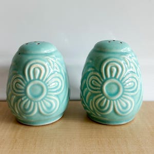 May include: A pair of turquoise ceramic salt and pepper shakers. Each shaker has a rounded, egg-like shape with a floral design. The floral pattern is raised and in a lighter shade of turquoise. The tops have three small holes for dispensing.