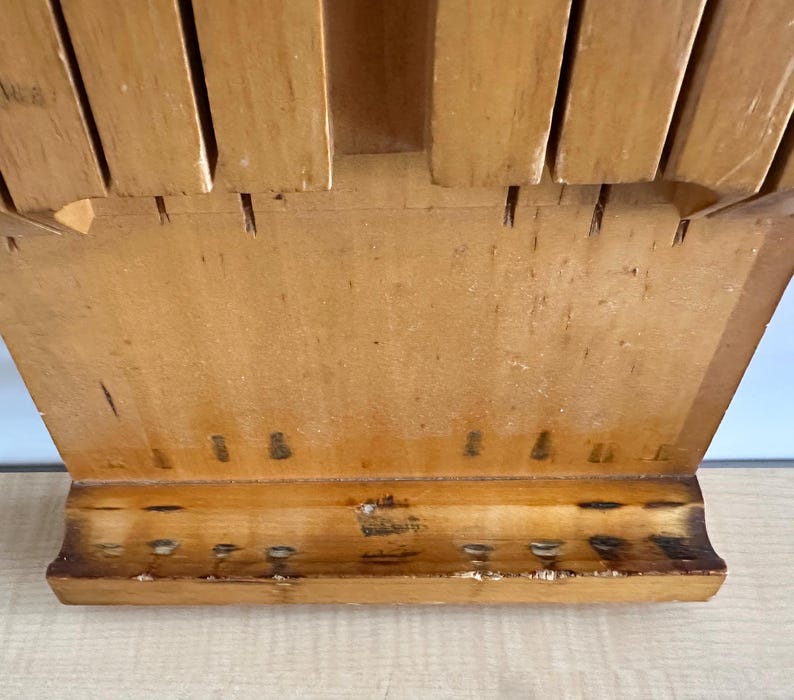 May include: A wooden knife block with slots for holding knives. The block is made of light brown wood and has a rectangular shape. The block has a small shelf at the bottom.