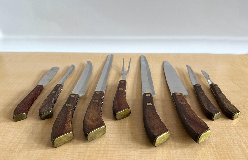 May include: Set of eight kitchen knives with wooden handles. The knives have stainless steel blades and brass accents. The knives are arranged on a wooden cutting board.