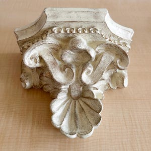 May include: A wall-mounted corbel featuring an off-white finish with distressed brown detailing. The architectural piece displays intricate scrollwork, a central shell motif at the base, and a decorative beaded border along the top edge.