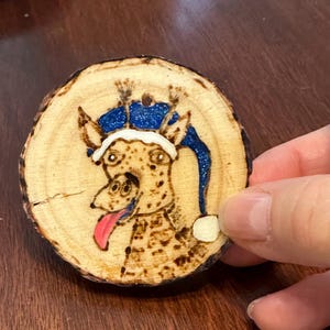 May include: A wood slice ornament featuring a whimsical giraffe design. The giraffe has a blue glittery hat and a pink tongue. The ornament is hand-burned with brown and black details, creating a rustic and unique piece.