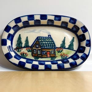 May include: A small, oval ceramic dish with a blue and white checkered border. The center of the dish features a hand-painted scene of a brown cabin with a red door, surrounded by green trees and red flowers.