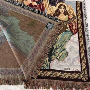 May include: A tapestry throw blanket with a woven design of angels playing musical instruments. The blanket has a brown, green, and gold color scheme with a fringe border. The text "OHMK. LIC., INC." is visible in the bottom right corner.