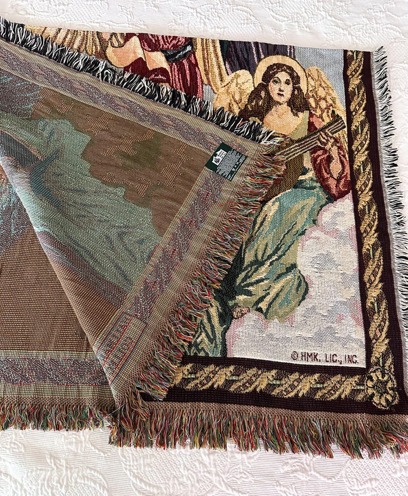 May include: A tapestry throw blanket with a woven design of angels playing musical instruments. The blanket has a brown, green, and gold color scheme with a fringe border. The text "OHMK. LIC., INC." is visible in the bottom right corner.