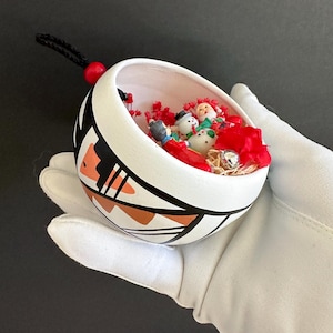 May include: A white ceramic ornament with a Southwestern design in black, orange, and brown. The ornament is filled with miniature Christmas figures and red decorative accents. A black cord with a red bead is attached for hanging.