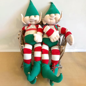 May include: Two plush green and red striped elves sitting on a wicker bench. The elves have green hats with bells, white faces, and red and white striped legs.