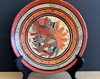 May include: A hand-painted ceramic plate with a vibrant, stylized bird design in the center. The plate features a red-orange border and a repeating geometric pattern. The plate is displayed on a black wooden stand.