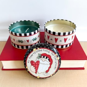 May include: Two round candles in decorative tins with a black and white checkered pattern, red hearts, and holly. A third tin with a Santa Claus illustration and the text "All hearts come home for Christmas" is in front.