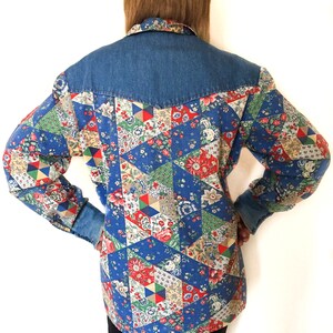 May include: A blue denim shirt with a patchwork design featuring a variety of floral patterns in red, white, and green. The shirt has a pointed collar and long sleeves.