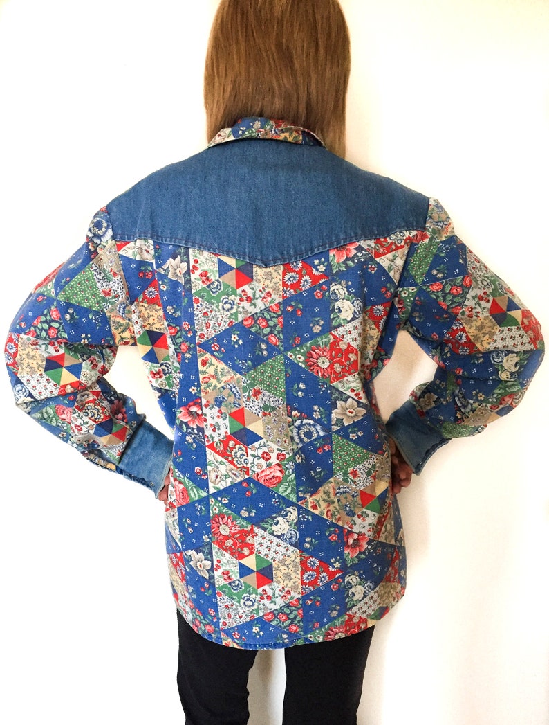 May include: A blue denim shirt with a patchwork design featuring a variety of floral patterns in red, white, and green. The shirt has a pointed collar and long sleeves.