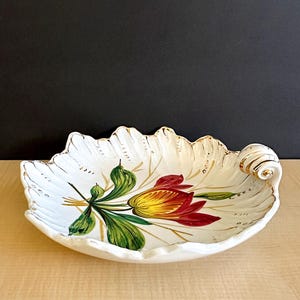 May include: White ceramic leaf-shaped dish with a hand-painted floral design. The dish features a red and yellow flower with green leaves and gold trim along the edges. The dish has a decorative swirl on one end.