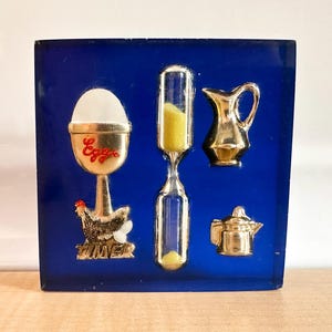 May include: A square, deep blue resin block featuring embedded miniature kitchen items: a gold-toned egg cup with a white egg and the red text "Eggs", a chicken figure with the text "TIMER", a glass hourglass with yellow sand, a small pitcher, and a teapot.
