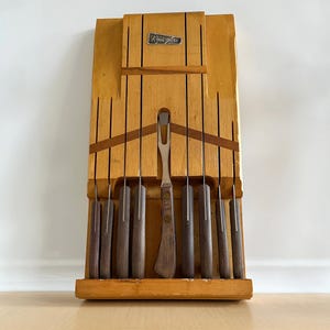 May include: A wooden knife block with a set of knives and a carving fork. The block is light brown and has a dark brown label that says "Kroma-fire".