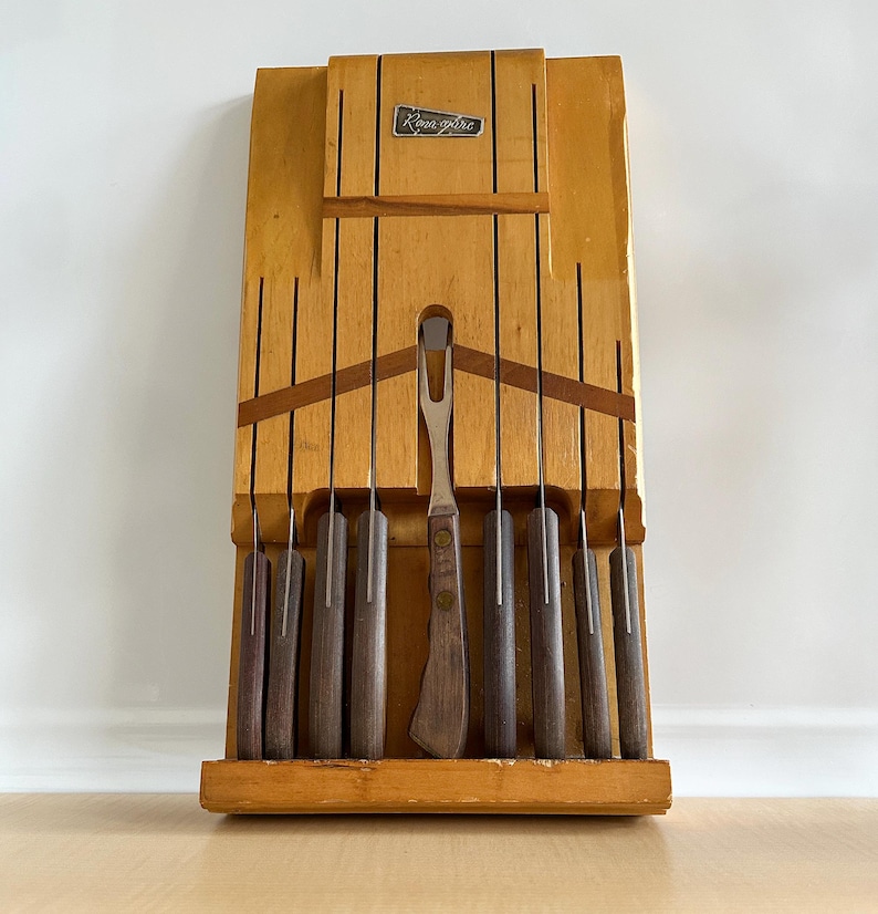 May include: A wooden knife block with a set of knives and a carving fork. The block is light brown and has a dark brown label that says "Kroma-fire".
