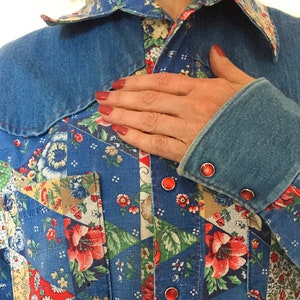 May include: A blue denim shirt with a patchwork design featuring floral patterns and red accents. The shirt has a pointed collar and a button-up closure. The sleeves are rolled up and have red buttons.