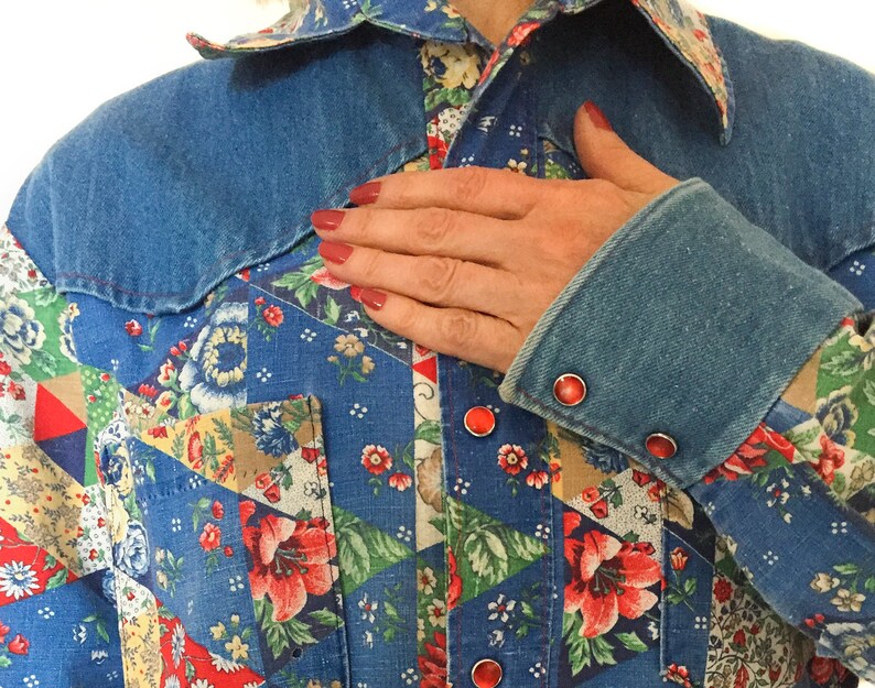May include: A blue denim shirt with a patchwork design featuring floral patterns and red accents. The shirt has a pointed collar and a button-up closure. The sleeves are rolled up and have red buttons.