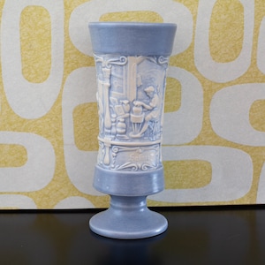 May include: A blue ceramic vase with white raised relief designs depicting a scene of people working in a bakery. The vase has a wide base and a narrow neck. The words "Salzburg" are written on the vase.