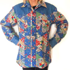 May include: A blue denim shirt with a patchwork design featuring floral patterns in shades of red, pink, yellow, and green. The shirt has a pointed collar and long sleeves.