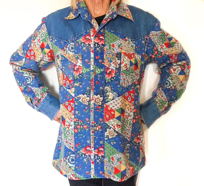 May include: A blue denim shirt with a patchwork design featuring floral patterns in shades of red, pink, yellow, and green. The shirt has a pointed collar and long sleeves.