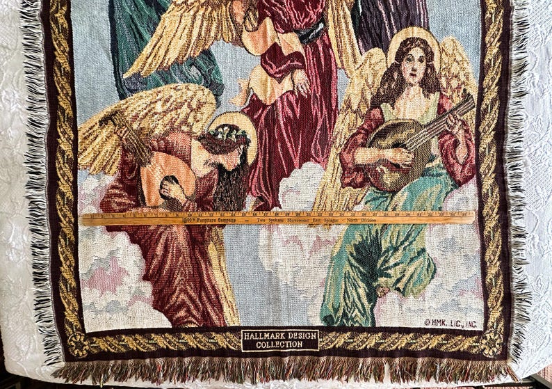 May include: A tapestry wall hanging featuring four angels with wings, two playing musical instruments. The tapestry has a brown and gold border with fringe. The text "Hallmark Design Collection" and "HMK, LLC" are visible at the bottom.