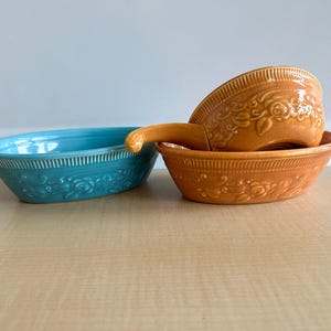 May include: Three ceramic serving dishes. One is oval and turquoise, the others are orange. The orange dishes are stacked, one with a handle. All dishes have a floral pattern and ribbed edges. The dishes are on a light wood surface.