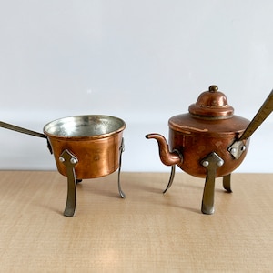 May include: Two miniature copper pots with handles and legs. The pots are on a wooden surface.