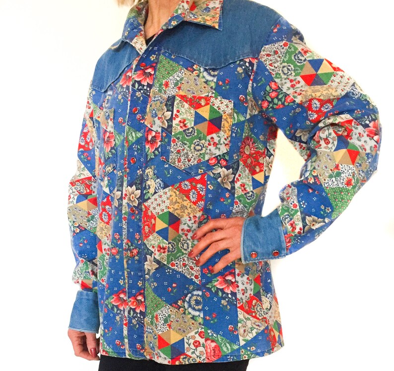May include: A blue denim shirt with a patchwork design featuring various floral patterns in red, yellow, green, and white. The shirt has a pointed collar and a button-up front.