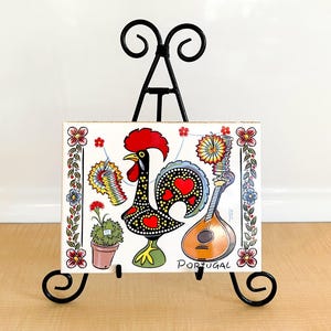 May include: A white ceramic tile with a colorful illustration of a rooster, a guitar, and flowers. The tile is on a black metal easel and says "Portugal" at the bottom.
