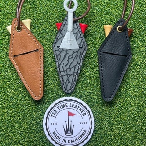 Puede incluir: Tres porta-tees de golf de cuero en marrón, gris y negro. Cada porta-tees tiene forma de diamante con una ranura para un tee. Los porta-tees están unidos a cordones de cuero marrón. Un círculo blanco con el texto "Tee Time Leather" y un logotipo de club de golf se encuentra en el centro inferior de la imagen.