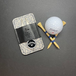 Op de afbeelding: Een grijze en zwarte leren golfbalmarker met een Callaway-logo en een zilveren divottool. Een witte golfbal met de tekst "Titleist 4" ligt naast twee houten golftees met blauwe strepen.