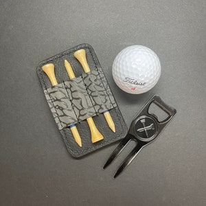 Op de afbeelding: Een zwarte leren golfteehouder met drie golftees, een zwarte golfbalretriever met een flesopener en een witte golfbal met de tekst "Titleist 4" erop gedrukt.