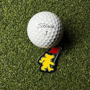 May include: A white Titleist golf ball with the number 3 and a yellow bear pin on a green surface. The pin has a red flag and black border. The golf ball has dimples.