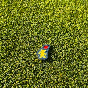 May include: A yellow and blue enamel pin with a red hat on top. The pin is shaped like a bear and is on a green background.