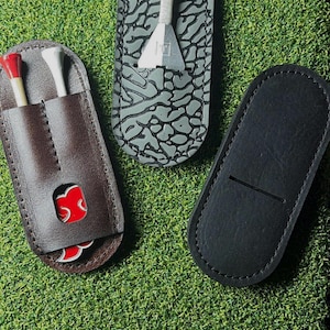 Puede incluir: Tres accesorios de golf sobre una superficie verde. Un soporte de tee de golf de cuero marrón con tees rojos y blancos, una herramienta de golf gris y negra y un soporte de marcador de bola de golf de cuero negro.