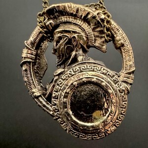 May include: A gold-toned pendant necklace featuring a detailed sculpture of a Roman soldier wearing a helmet and holding a shield. The shield is decorated with a Greek key pattern.