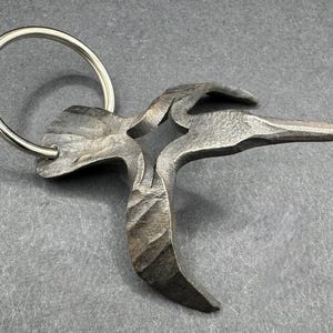 May include: A hand-forged metal keyring with a bird-like design. The keyring has a textured, dark grey finish and a silver-coloured ring. The bird's form is abstract, with a long beak and outstretched wings. The item is likely made of iron.