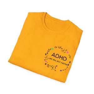Op de afbeelding: Mosterdgeel T-shirt met een ronde hals. Het shirt heeft een zwart cirkelvormig ontwerp met het woord "ADHD" en de zin "LIKE YOU, BUT FASTER" in een speels lettertype. Het ontwerp bevat kleurrijke harten, sterren en andere vormen.