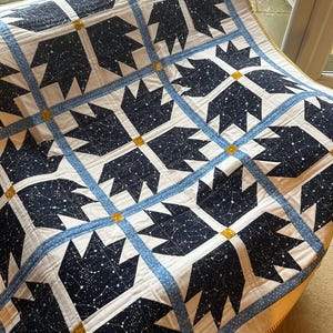 May include: A handmade quilt with a repeating pattern of dark blue bear paw shapes filled with a constellation design on a white background. The quilt is bordered with light blue and gold accents, creating a cosy, celestial theme.