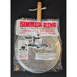 Könnte beinhalten: Ein Vintage-Simmerring-Hitzediffusor für Gas- und Elektroherde. Die silberfarbene, perforierte Scheibe hat einen Holzgriff und ist in einer transparenten Plastiktüte verpackt. Der Text auf der Verpackung lautet "SIMMER RING" und enthält Gebrauchsanweisungen.