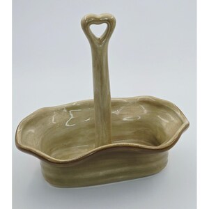 May include: A beige ceramic serving dish with a heart-shaped handle. The dish has a wavy rim and a brown border. The handle is centered and extends upwards from the center of the dish.